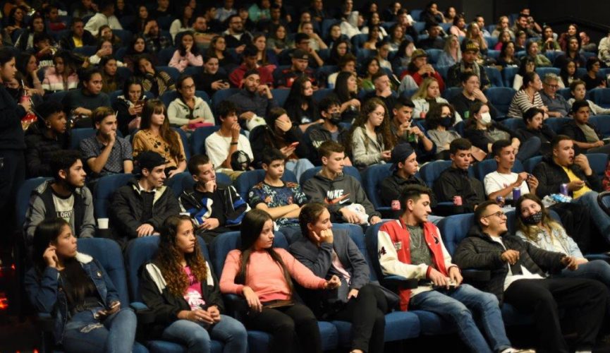 Adolecentes sentados em cadeiras de cinema. Foto ampla, mostrando muitas pessoas.