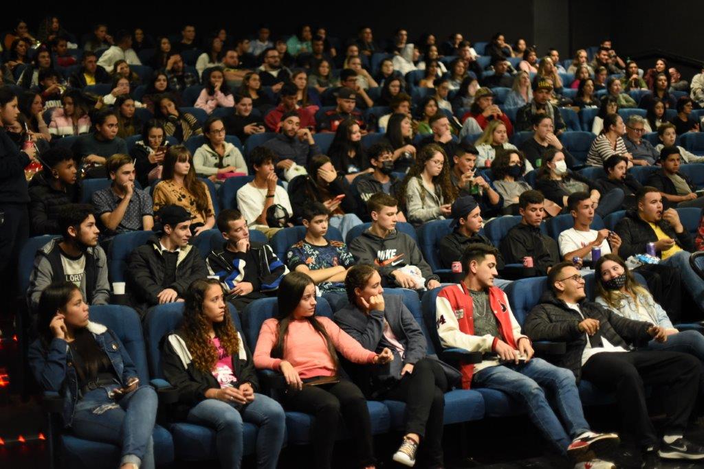 Adolecentes sentados em cadeiras de cinema. Foto ampla, mostrando muitas pessoas.